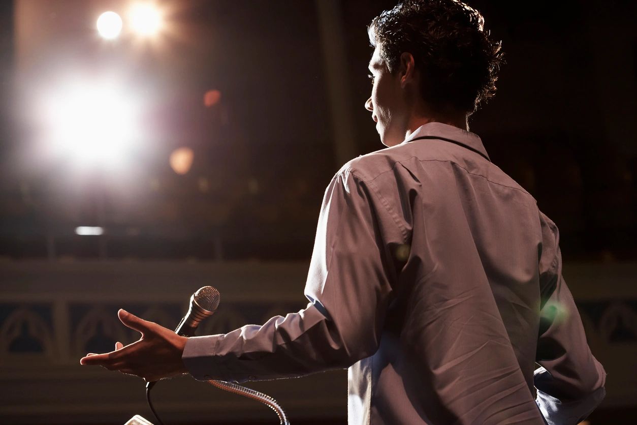 A speaker addressing an audience with a microphone, seen from behind.